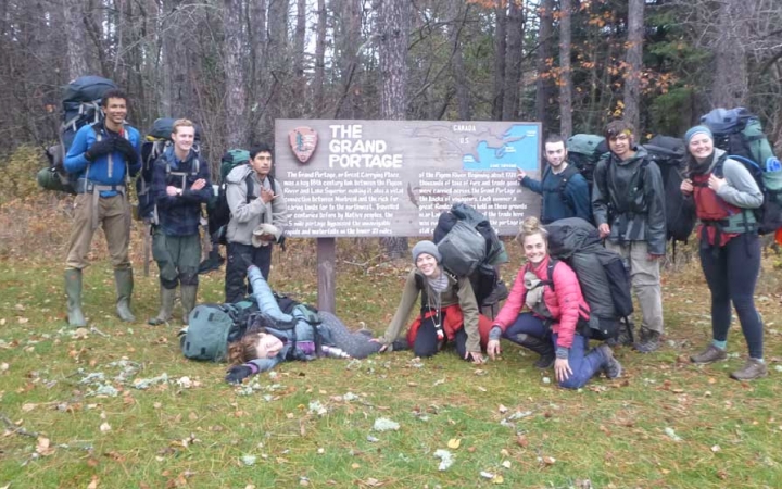 A group of students wearing backpacks pose for a photo with a sign that reads "The Grand Portage," with additional writing, too small to read. 